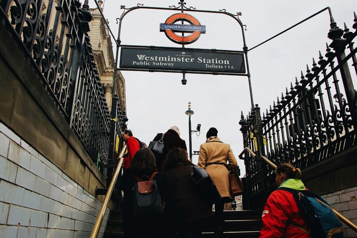 London 2026 travel reality: a tired traveler exiting a Tube station into fast pedestrian flow at a busy junction while checking directions