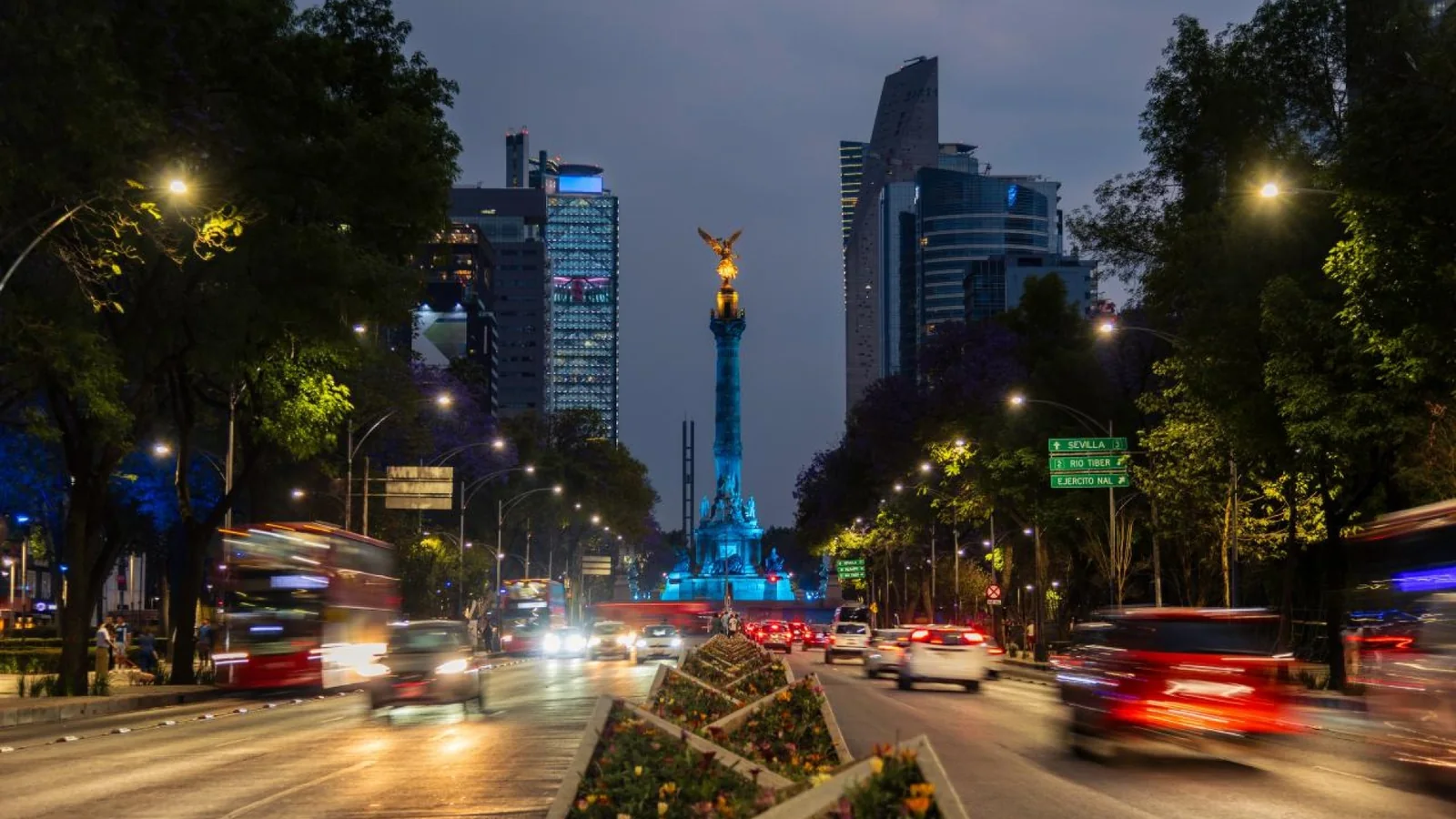 The Angel of Independence illuminated at night in Mexico City with traffic and skyscrapers surrounding the landmark