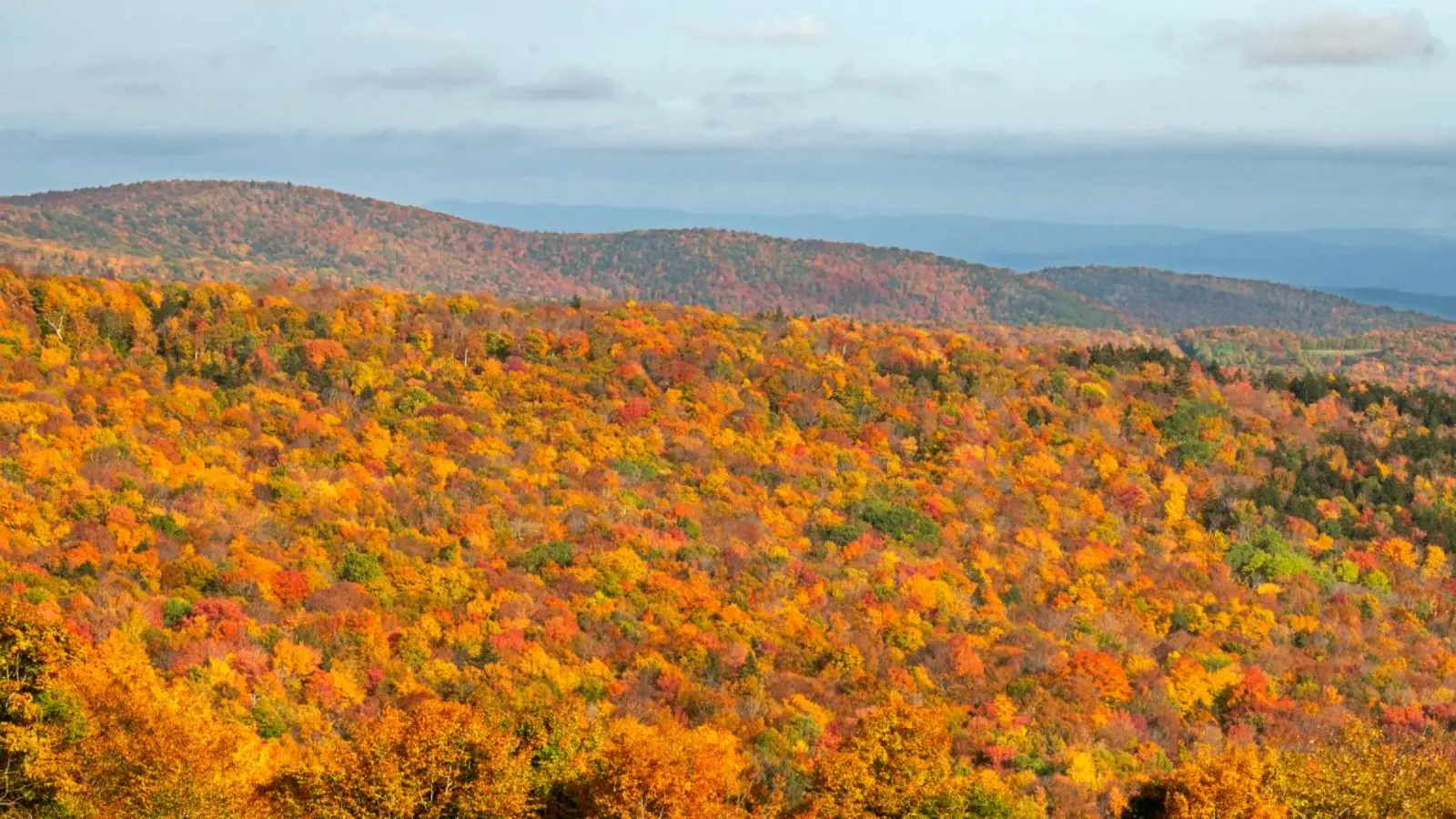 Rolling hills beneath distant mountain ridges display fall foliage in Vermont.