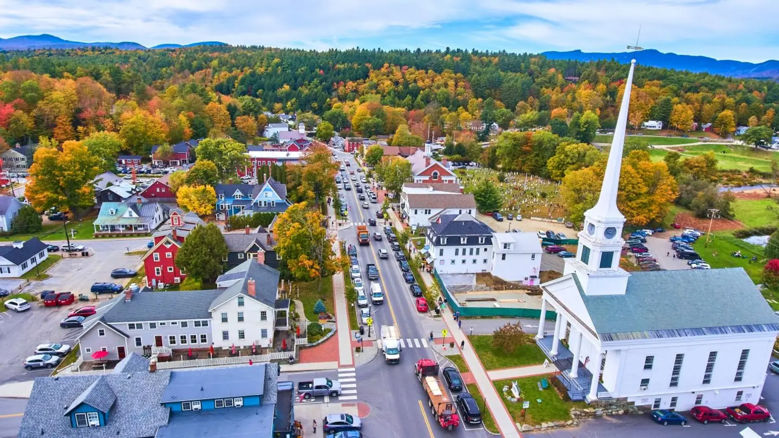 Aerial view of a village street with autumn trees, parked cars, and a white church steeple.