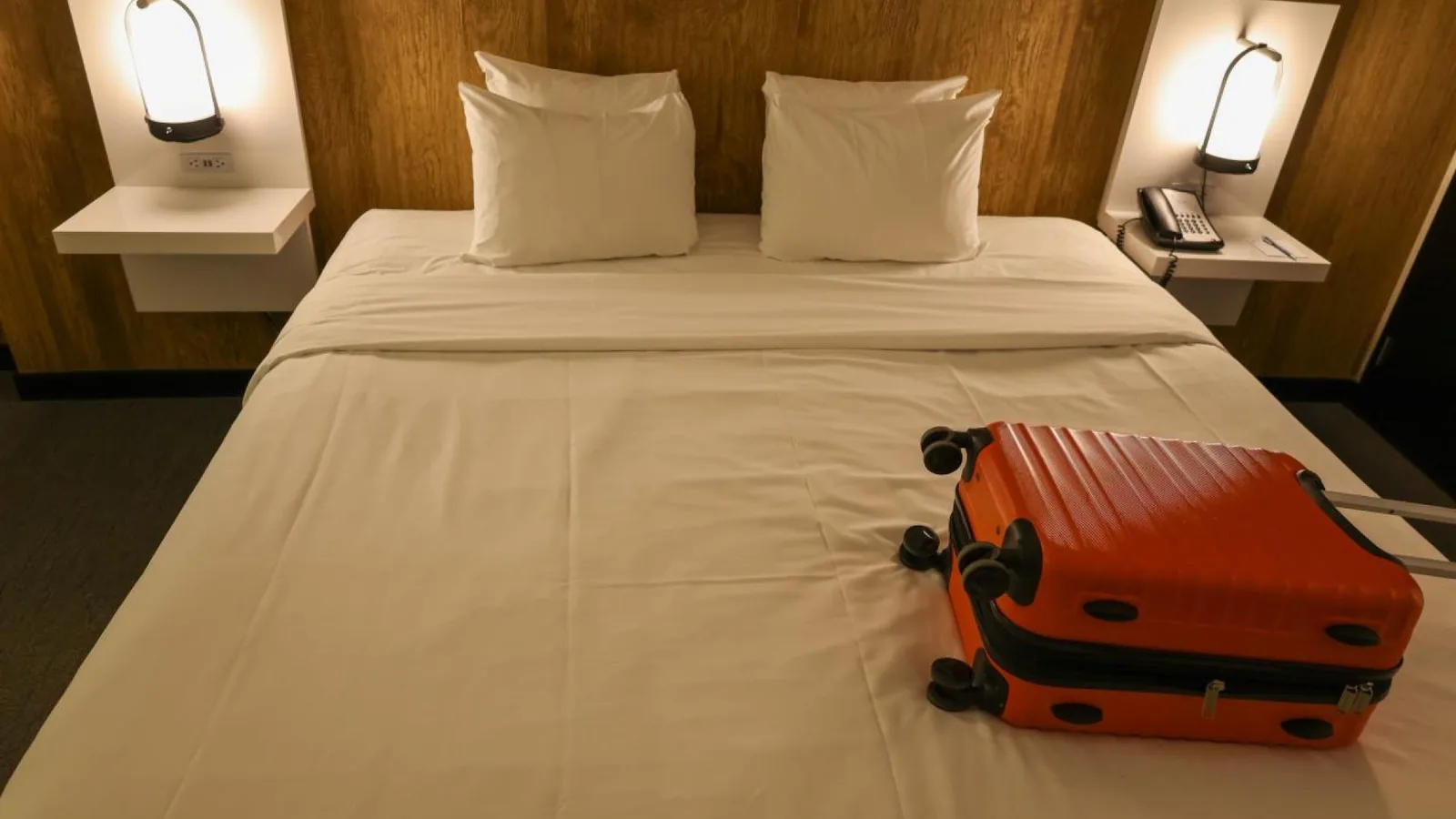 Orange suitcase on a white hotel bed with bedside lamps and a wooden headboard