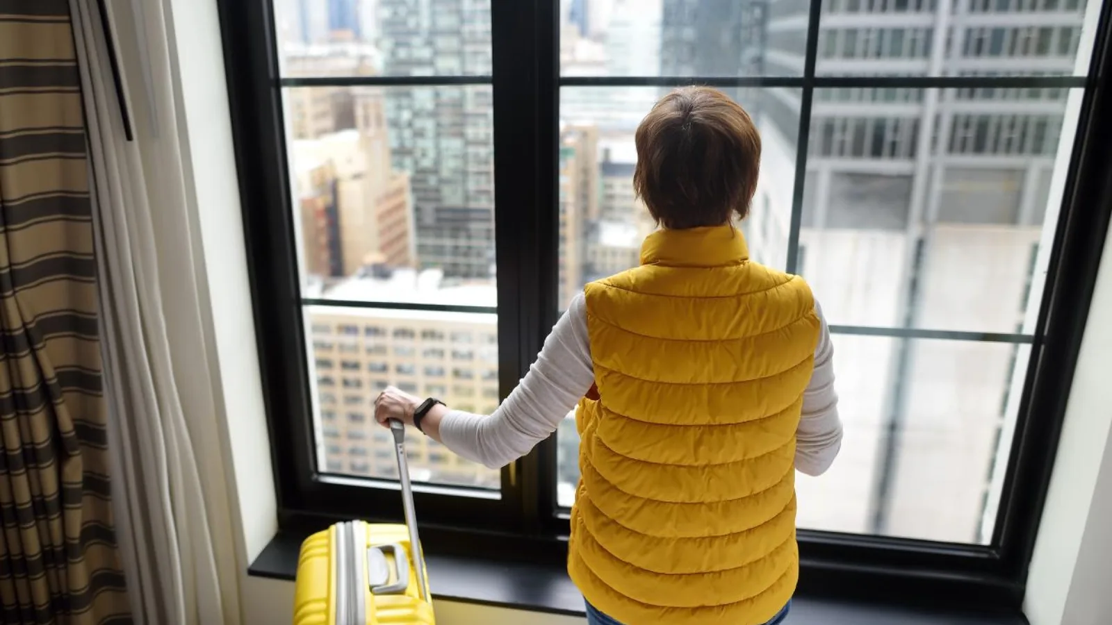 Traveler with a yellow suitcase looking out of a large window toward city buildings