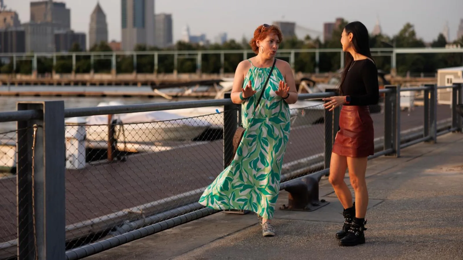 Two women talking beside a waterfront railing, with boats and city buildings in the background